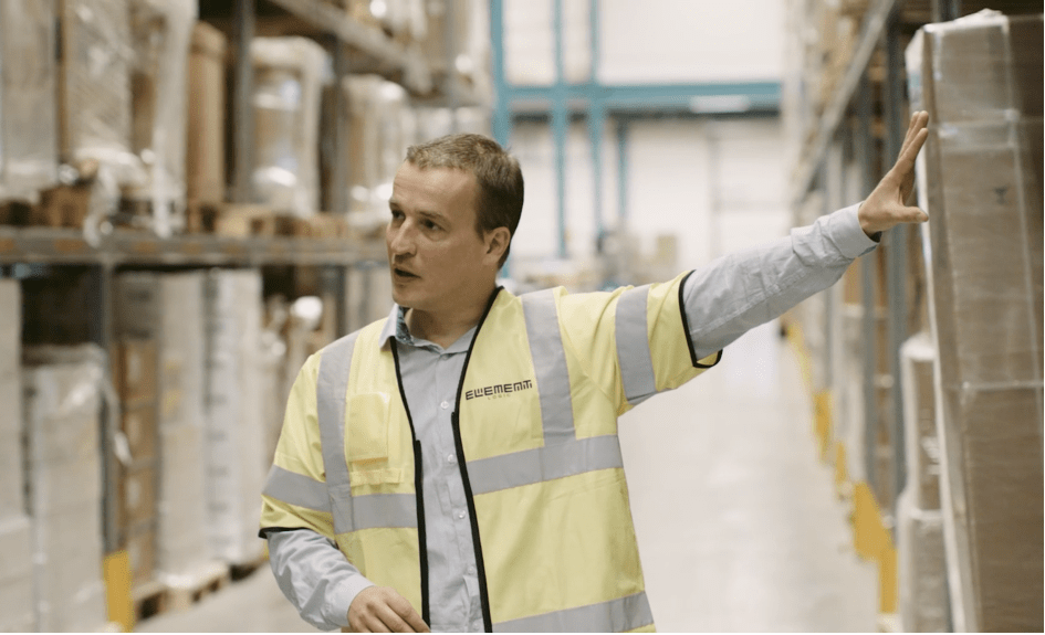 A man in a yellow west in a warehouse is pointing and looking at something outside of the camera shot.