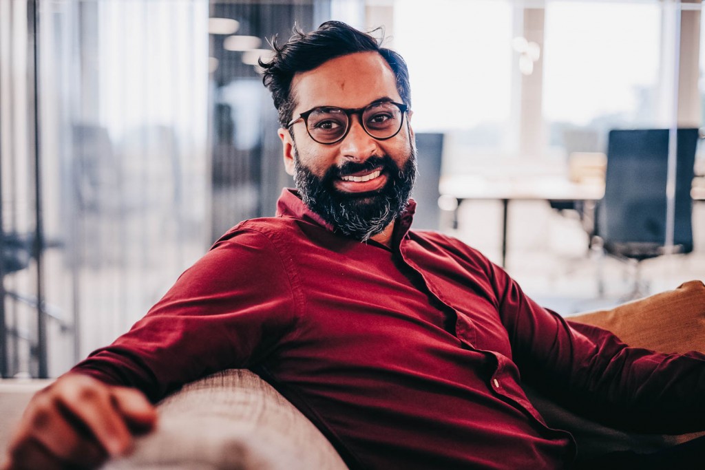 A smiling Rahman sits om a couch while looking into the camera.